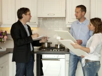 A designer discusses cabinet door options with a couple in a modern white kitchen.